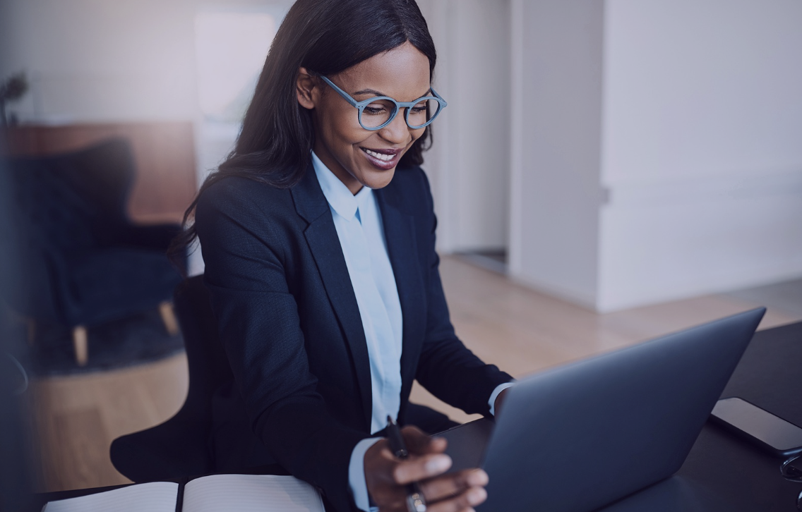 Professional woman wearing glasses and a navy business suit smiling while working on a laptop at a desk, with a notebook and phone beside her.