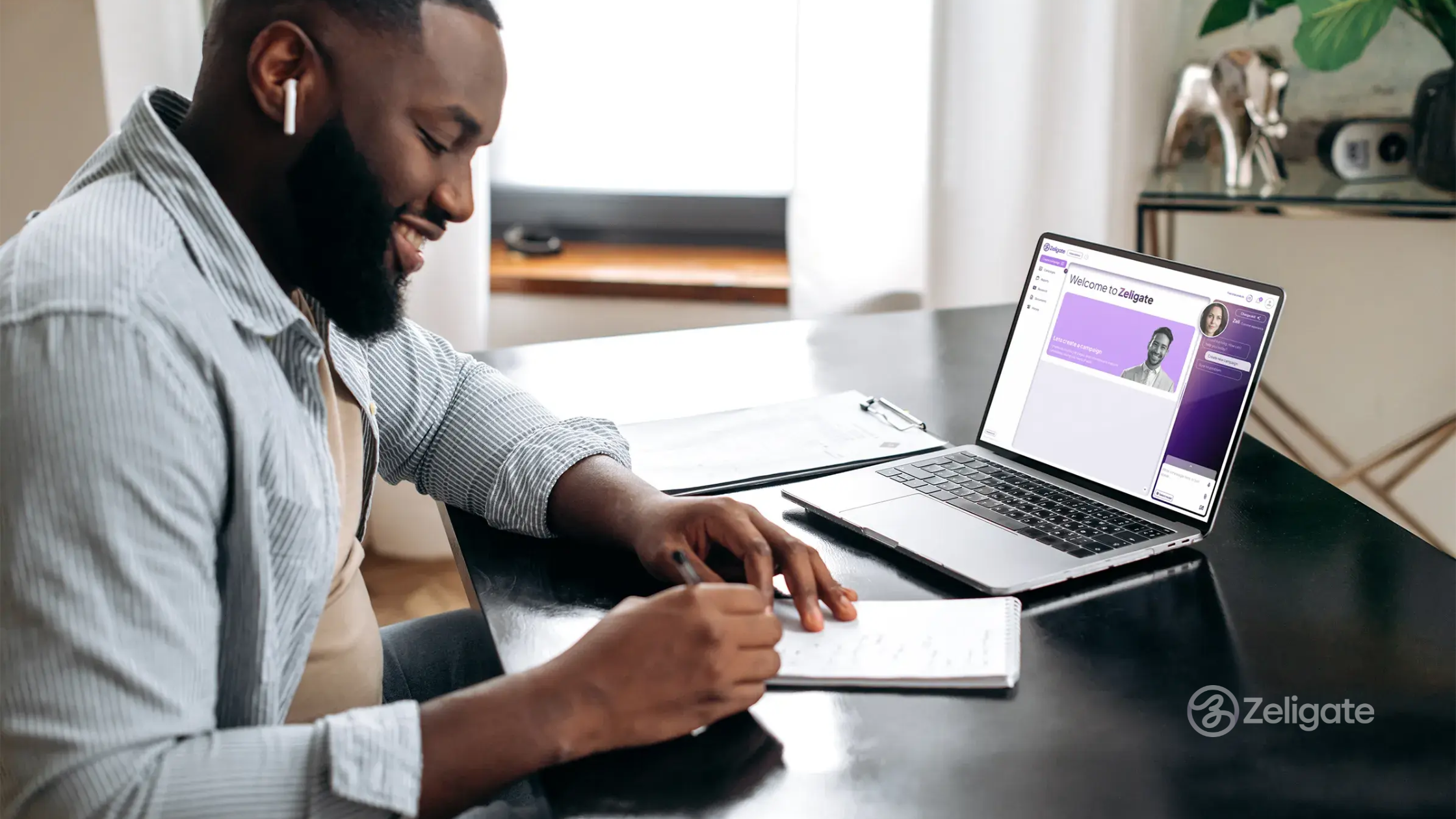 A man wearing wireless earbuds smiles while taking notes at his desk. He’s engaged with Zeligate’s AI assistant on his laptop, which displays a hiring support interface. The setting is a bright, modern home office.