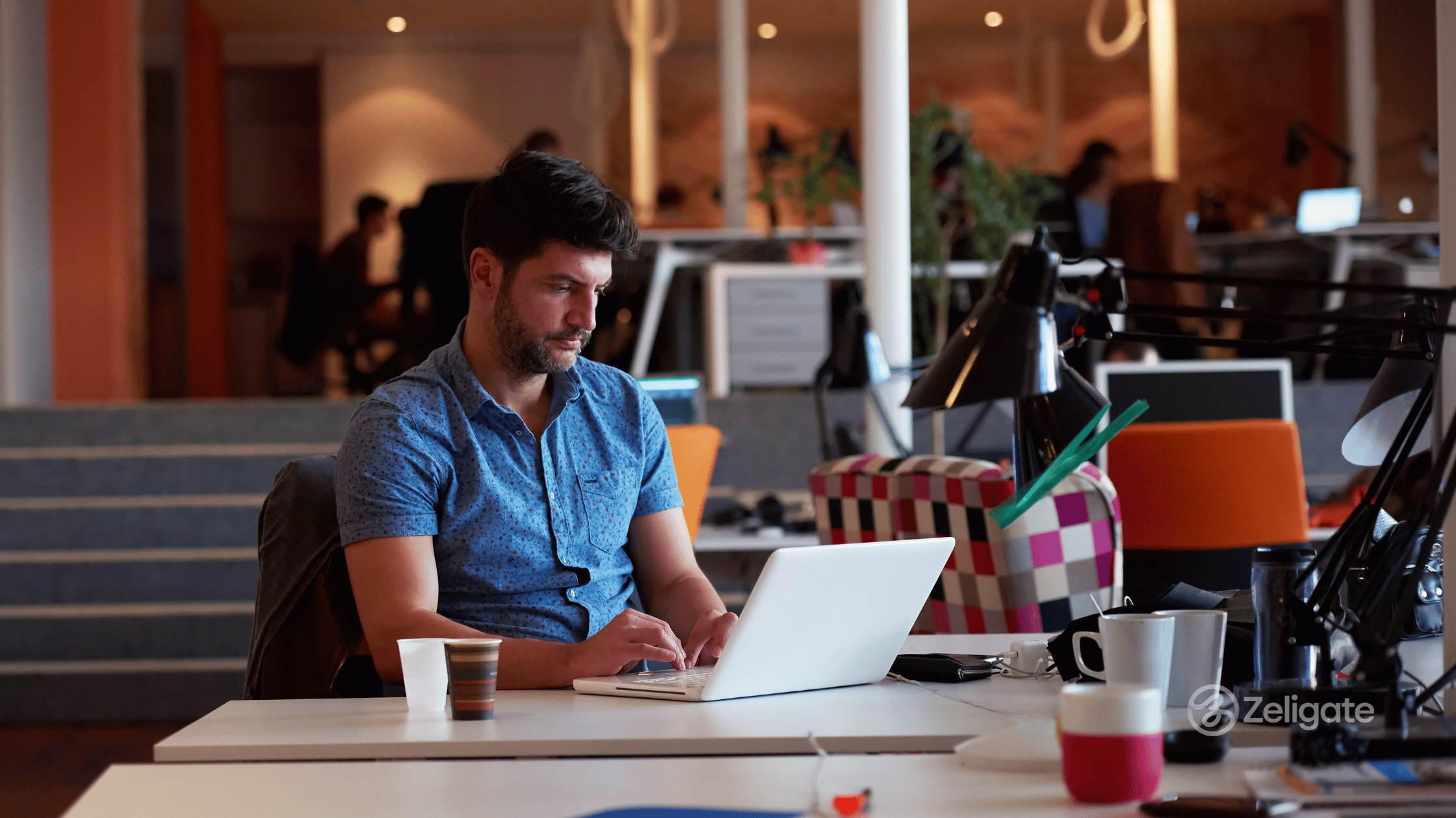 A man working on a laptop in a modern open-plan office, representing startup hiring and productivity, with the Zeligate logo in the corner.