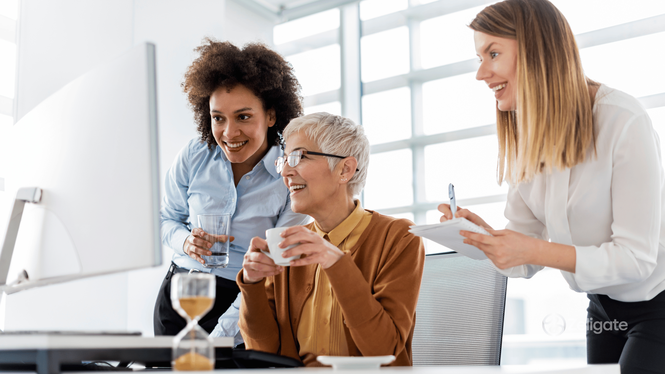 Three diverse professionals collaborating around a computer in a bright office, smiling and discussing recruitment workflows, illustrating teamwork and AI-driven hiring efficiency.